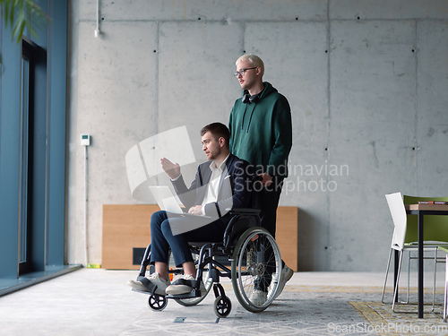 Image of A businessman in a wheelchair in a fashionable office using a laptop while behind him is his business colleague who gives him support