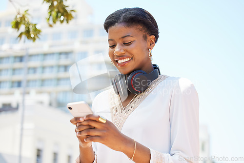 Image of Phone, city and music with a business black woman typing a text message while walking to work outdoor. Mobile, headphones and social media with a happy employee commuting in an urban town with flare