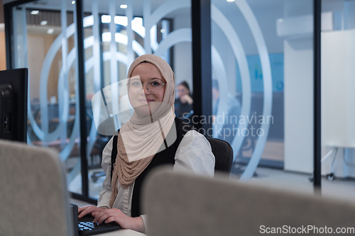 Image of In a modern office, a young Muslim entrepreneur wearing a hijab sits confidently and diligently works on her computer, embodying determination, creativity, and empowerment in the business world