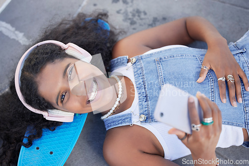 Image of Portrait, phone and music with a black woman skater lying on her board at a skatepark from above. Face, skating and social media with a female skateboarder resting on the ground during a break