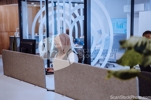 Image of In a modern office, a young Muslim entrepreneur wearing a hijab sits confidently and diligently works on her computer, embodying determination, creativity, and empowerment in the business world