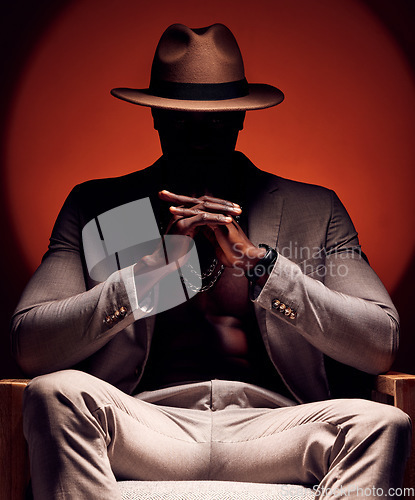 Image of Stylish, fashionable and trendy man in silhouette sitting in a dark room with a red studio background. Edgy, mysterious and curious male mafia boss sitting in a fancy suit, hat and expensive jewelry