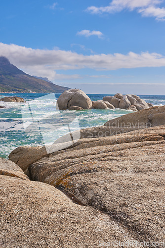 Image of Beautiful sea view of a big boulders and ocean water on a sunny beach day in summer. A seaside setting in nature with a blue sky, white clouds, and waves. A seascape near a shore and mountains.