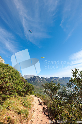 Image of Nature park landscape with plants and an outdoor dirt path. Beautiful outdoors view of a hiking trail in the mountains during summer. Mountain background with a blue sky and paragliding travelers.