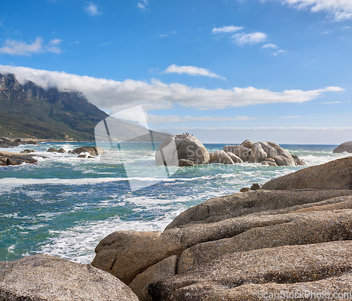 Image of A calming ocean view of the rocky coast of Camps Bay in Cape Town, South Africa. Boulders at a beach with the tide and current coming in and Table Mountain on the horizon. Tidal waves washing ashore