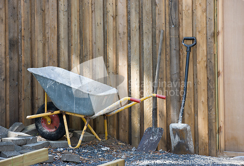 Image of A wheelbarrow and spade placed amongst gravel and building material. Construction site at a home in the backyard. Equipment and tools used to build a house. A shovel and other building equipment