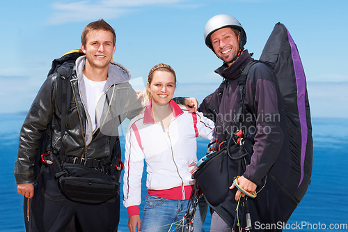 Image of Men, woman and portrait with happy for paragliding, extreme sport and outdoor by sea in sunshine. Group, fitness and smile on adventure with helmet, backpack or parachute for jump, safety or blue sky