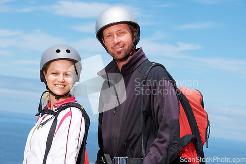 Image of Man, woman and portrait with smile for paragliding, extreme sport and outdoor by sea in sunshine. Happy couple, fitness and adventure with helmet, backpack or parachute for jump, safety and blue sky