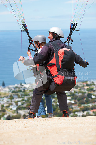 Image of People, paragliding and outdoor to land on ground, extreme sport and sunshine by ocean on hill. Coach, person and back on adventure with helmet, backpack or parachute for jump, safety and blue sky