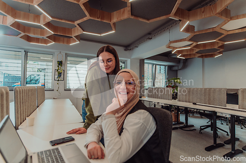 Image of Young businesswomens, one of them wearing a hijab, are collaboratively problem-solving in a modern office while working on a laptop, exemplifying diversity, professionalism, and empowered teamwork