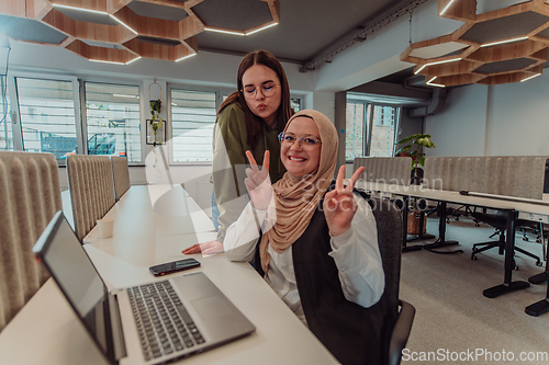 Image of Young businesswomens, one of them wearing a hijab, are collaboratively problem-solving in a modern office while working on a laptop, exemplifying diversity, professionalism, and empowered teamwork