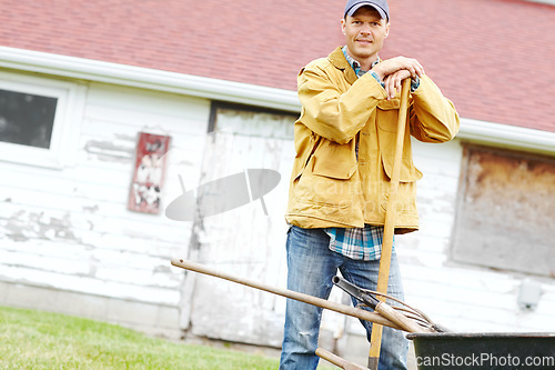 Image of Manual labour. Portrait of a happy man standing next to a wheelbarrow with a gardening tool in his hand.