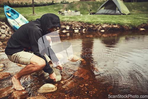 Image of Nature has everything you need. a young man filling water from a lake into a pot at a campsite.