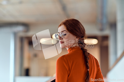 Image of In a vibrant modern startup office, a businesswoman with striking orange hair is immersed in her work at her desk, embodying the dynamic and creative spirit of contemporary entrepreneurship.