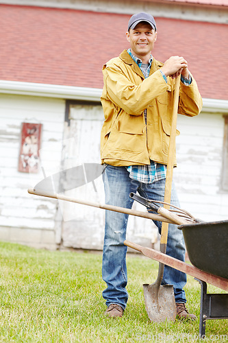 Image of He works hard for his money. Portrait of a happy man standing next to a wheelbarrow with a gardening tool in his hand.