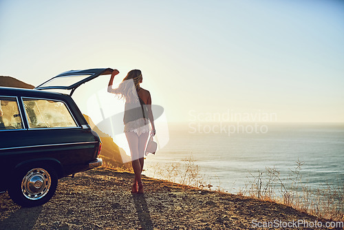 Image of Its a view worth driving for. Rearview shot of a young woman standing next to her car during a roadtrip.