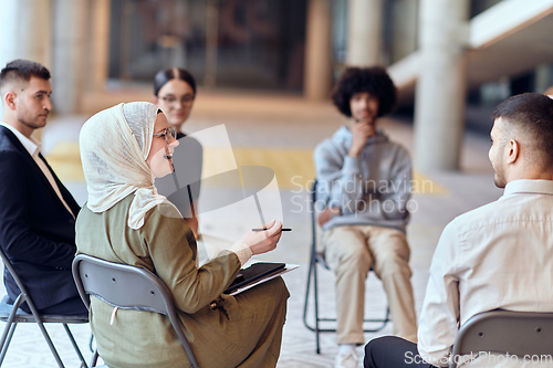 Image of A diverse group of young business entrepreneurs gathered in a circle for a meeting, discussing corporate challenges and innovative solutions within the modern confines of a large corporation