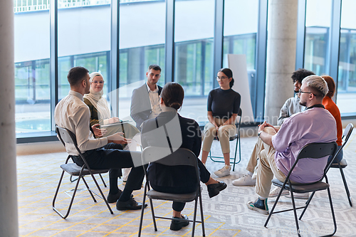 Image of A diverse group of young business entrepreneurs gathered in a circle for a meeting, discussing corporate challenges and innovative solutions within the modern confines of a large corporation