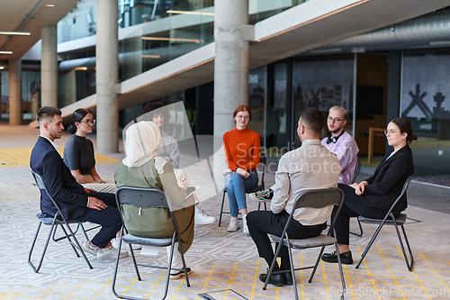 Image of A diverse group of young business entrepreneurs gathered in a circle for a meeting, discussing corporate challenges and innovative solutions within the modern confines of a large corporation