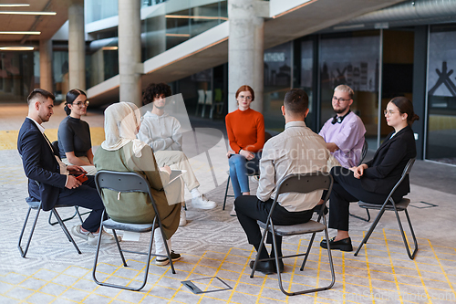 Image of A diverse group of young business entrepreneurs gathered in a circle for a meeting, discussing corporate challenges and innovative solutions within the modern confines of a large corporation