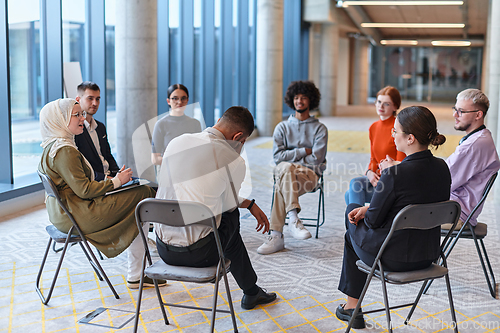 Image of A diverse group of young business entrepreneurs gathered in a circle for a meeting, discussing corporate challenges and innovative solutions within the modern confines of a large corporation