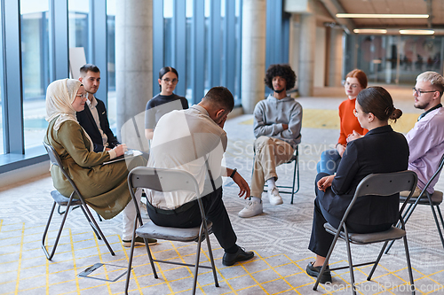 Image of A diverse group of young business entrepreneurs gathered in a circle for a meeting, discussing corporate challenges and innovative solutions within the modern confines of a large corporation