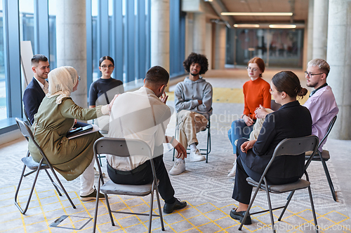 Image of A diverse group of young business entrepreneurs gathered in a circle for a meeting, discussing corporate challenges and innovative solutions within the modern confines of a large corporation