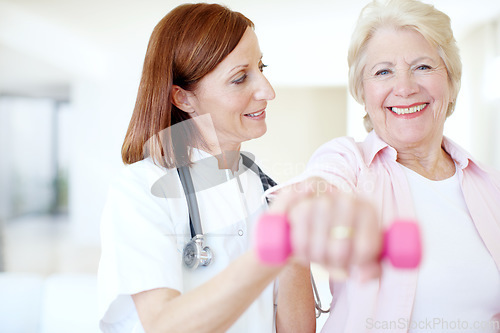 Image of The strength of her resolve - Senior Care. Smiling elderly female is assisted by her nurse as she lifts a dumbbell - Focus on background.