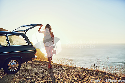 Image of On the hunt for wild open spaces. Rearview shot of a young woman standing next to her car during a roadtrip.