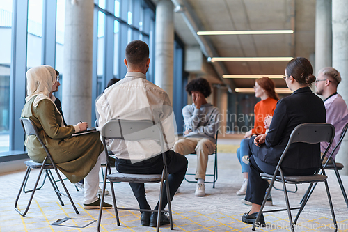 Image of A diverse group of young business entrepreneurs gathered in a circle for a meeting, discussing corporate challenges and innovative solutions within the modern confines of a large corporation