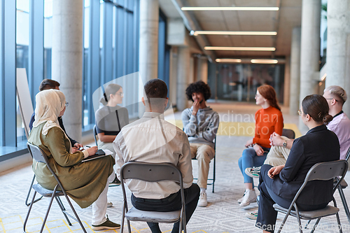 Image of A diverse group of young business entrepreneurs gathered in a circle for a meeting, discussing corporate challenges and innovative solutions within the modern confines of a large corporation