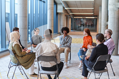 Image of A diverse group of young business entrepreneurs gathered in a circle for a meeting, discussing corporate challenges and innovative solutions within the modern confines of a large corporation