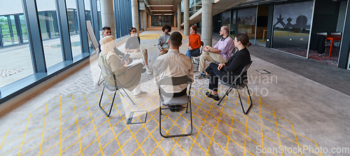 Image of A diverse group of young business entrepreneurs gathered in a circle for a meeting, discussing corporate challenges and innovative solutions within the modern confines of a large corporation