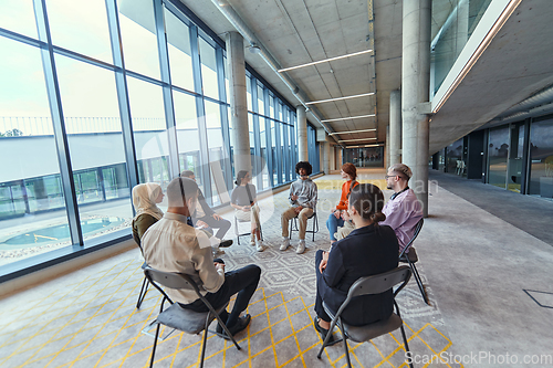 Image of A diverse group of young business entrepreneurs gathered in a circle for a meeting, discussing corporate challenges and innovative solutions within the modern confines of a large corporation