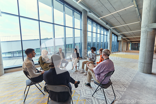 Image of A diverse group of young business entrepreneurs gathered in a circle for a meeting, discussing corporate challenges and innovative solutions within the modern confines of a large corporation