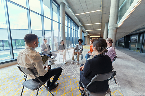 Image of A diverse group of young business entrepreneurs gathered in a circle for a meeting, discussing corporate challenges and innovative solutions within the modern confines of a large corporation