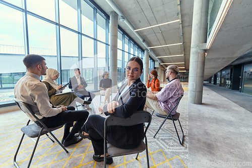 Image of A diverse group of young business entrepreneurs gathered in a circle for a meeting, discussing corporate challenges and innovative solutions within the modern confines of a large corporation