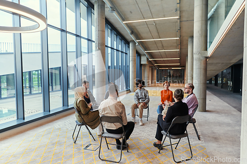 Image of A diverse group of young business entrepreneurs gathered in a circle for a meeting, discussing corporate challenges and innovative solutions within the modern confines of a large corporation