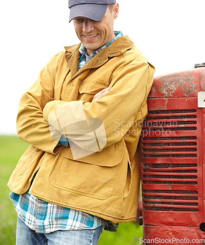 Image of Living a good and simple life. a smiling farmer standing next to his tractor with his arms crossed and looking down.