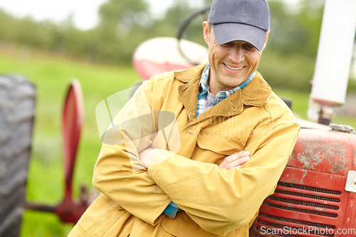 Image of Hes happy with his farm. a smiling farmer standing next to his tractor with his arms crossed and looking down.