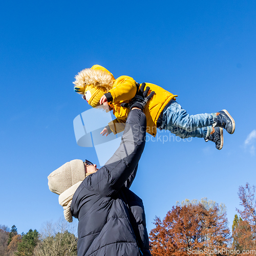 Image of More, more,...mum, that's fun. Happy young mother throws her cute little baby boy up in the air. Mother's Day, Mather and her son baby boy playing and hugging outdoors in nature in fall.