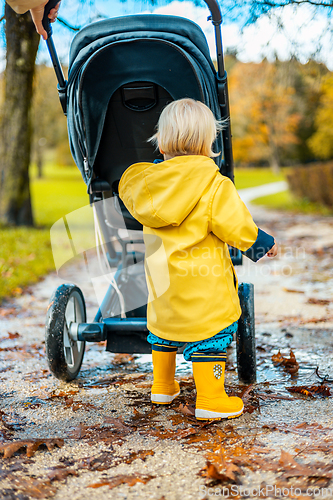 Image of Sun always shines after the rain. Small blond infant boy wearing yellow rubber boots and yellow waterproof raincoat walking in puddles, pushing stroller in city park, holding mother's hand after rain.