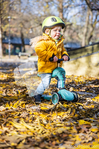 Image of Adorable toddler boy wearing yellow protective helmet riding baby scooter outdoors on autumn day. Kid training balance on mini bike in city park. Fun autumn outdoor activity for small kids.