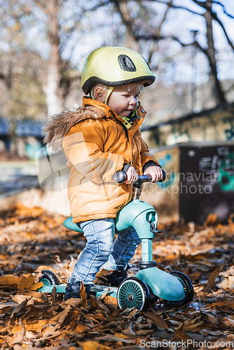 Image of Adorable toddler boy wearing yellow protective helmet riding baby scooter outdoors on autumn day. Kid training balance on mini bike in city park. Fun autumn outdoor activity for small kids.