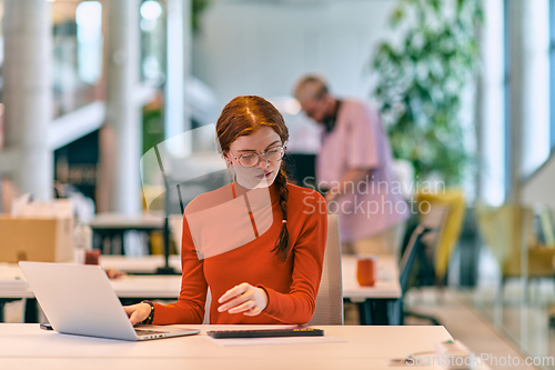 Image of In a modern startup office, a professional businesswoman with orange hair sitting at her laptop, epitomizing innovation and productivity in her contemporary workspace.