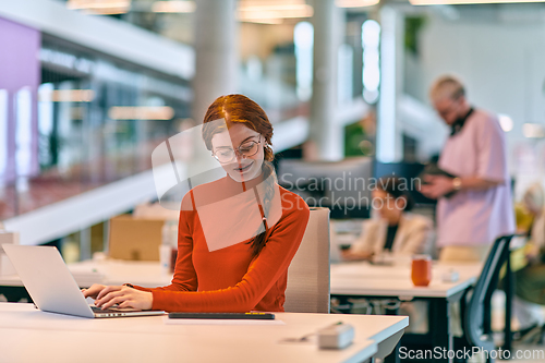 Image of In a modern startup office, a professional businesswoman with orange hair sitting at her laptop, epitomizing innovation and productivity in her contemporary workspace.