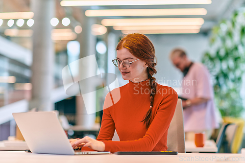 Image of In a modern startup office, a professional businesswoman with orange hair sitting at her laptop, epitomizing innovation and productivity in her contemporary workspace.