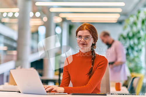 Image of In a modern startup office, a professional businesswoman with orange hair sitting at her laptop, epitomizing innovation and productivity in her contemporary workspace.