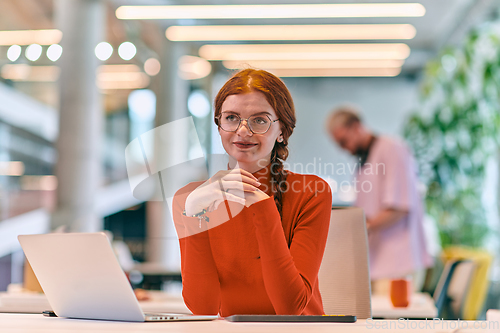 Image of In a modern startup office, a professional businesswoman with orange hair sitting at her laptop, epitomizing innovation and productivity in her contemporary workspace.