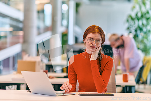 Image of In a modern startup office, a professional businesswoman with orange hair sitting at her laptop, epitomizing innovation and productivity in her contemporary workspace.